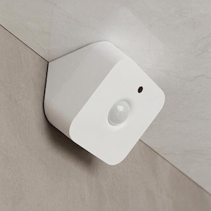 Op de afbeelding: Een witte bewegingssensor met een geometrisch ontwerp, gemonteerd in een hoek. Het heeft een afgeronde lens en een kleine zwarte stip. De sensor is geïnstalleerd op een lichtgrijze betegelde ondergrond.