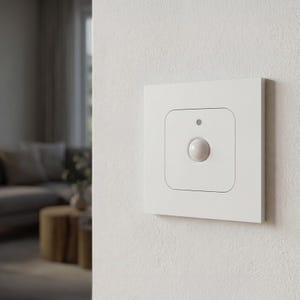 Op de afbeelding: Een wit, vierkant smart home-apparaat bevestigd aan een witte, getextureerde muur. Het apparaat heeft een centrale, ronde knop en een kleine stip erboven. De achtergrond toont een vage woonkamer.