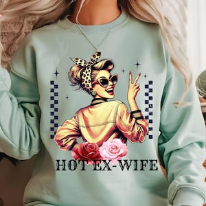 May include: A pale green sweatshirt with a vintage-style illustration of a woman in sunglasses and a headscarf, making a peace sign. The design includes the text "HOT EX-WIFE" and roses.