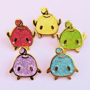May include: Five colorful enamel pins featuring a cute cartoon character design. Each pin has a gold outline and is filled with glitter in red, yellow, green, purple, and blue. The pins have a rounded body, small arms and legs, and a small antenna.