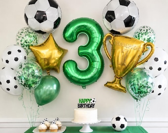 May include: A soccer-themed party decoration set featuring a large green number 3 balloon, a gold trophy balloon with the text CHAMPION, two soccer ball balloons, a gold star balloon, green confetti balloons, and a cake topper reading HAPPY BIRTHDAY.