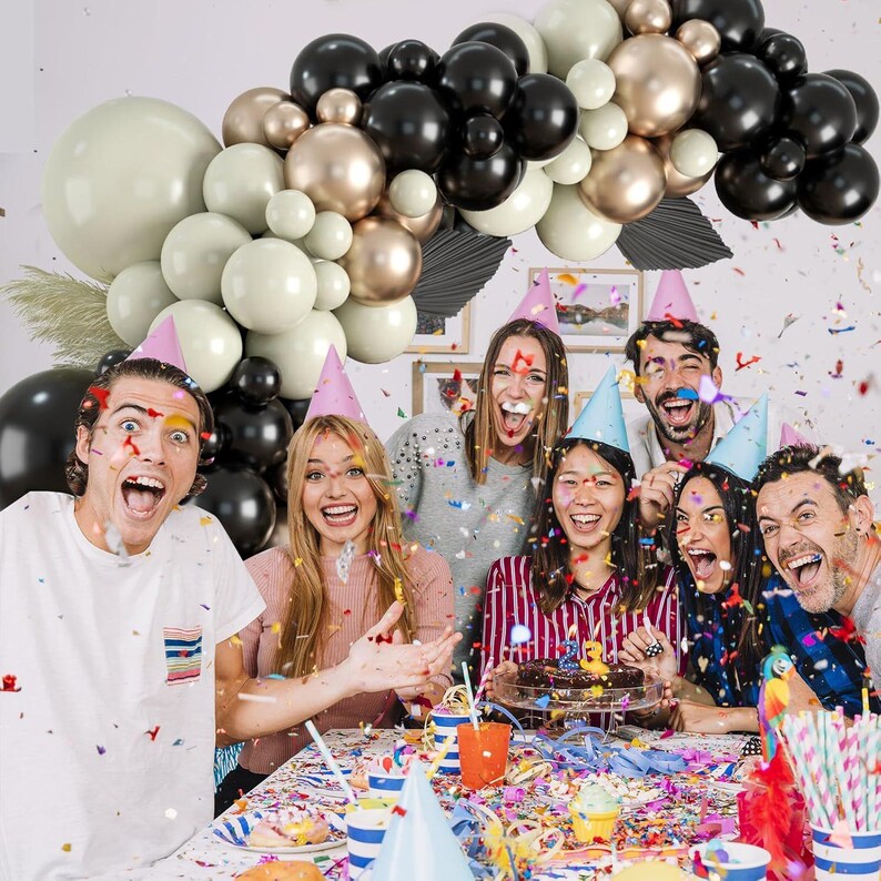 May include: A group of friends celebrating with a birthday cake, confetti, and a balloon arch made of black, gold, and white balloons.