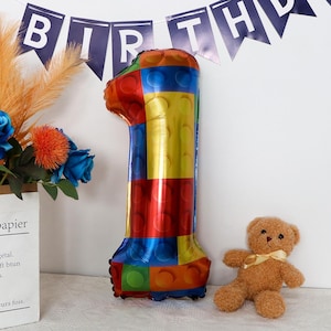 May include: A large, colorful, foil balloon shaped like the number one. The balloon is decorated with a pattern of red, yellow, blue, and green squares, resembling building blocks. The balloon is standing on a white surface.