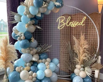 May include: A white metal arch with a grid pattern decorated with a balloon garland in shades of blue and beige. The garland is made of various sizes of balloons and includes a neon sign that reads "Blessed".