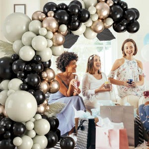 May include: A black, white, and gold balloon arch with three women standing in front of it. The women are smiling and holding drinks. The balloons are arranged in a circular shape and are tied together with string.