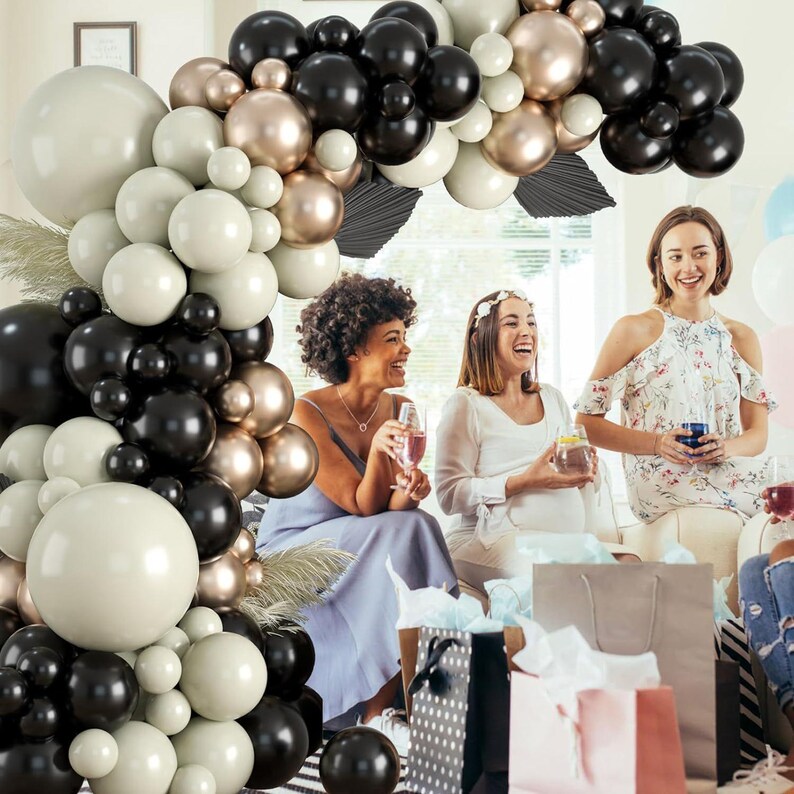 May include: A black, white, and gold balloon arch with three women standing in front of it. The women are smiling and holding drinks. The balloons are arranged in a circular shape and are tied together with string.