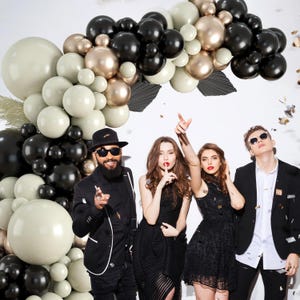 May include: Four people pose in front of a balloon arch made of black, white, and gold balloons. The arch is in the shape of a half-circle. The people are wearing black and white clothing.