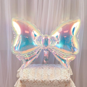 Op de afbeelding: Een grote, regenboogkleurige folie ballon in de vorm van een strik. De ballon is lichtblauw met een regenboogkleurige glans.