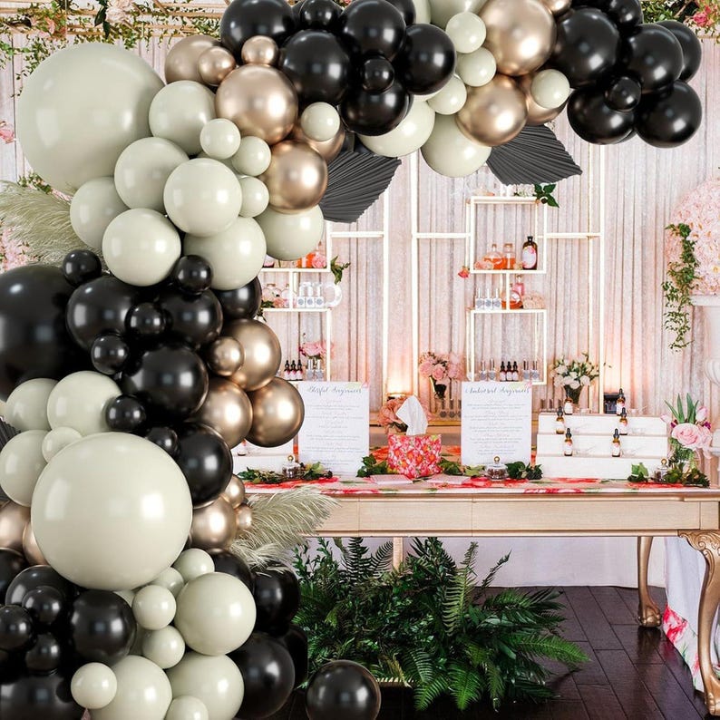 May include: A balloon arch made of black, gold, and cream balloons. The arch is decorated with greenery and is set up in front of a white wall with shelves.