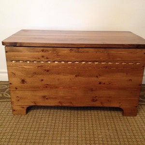 May include: A wooden storage chest with a hinged lid. The chest is made from light brown wood with a darker brown lid. A decorative row of carved details runs across the front. The chest sits on short, curved feet.