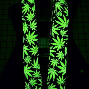 May include: A black guitar strap with a repeating pattern of bright green cannabis leaves. The strap is illuminated, creating a glowing effect against the dark background. The strap has two end pieces with a shield shape.