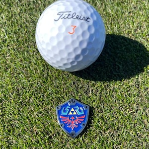 May include: A white Titleist golf ball with the number 3 in orange, next to a blue, red, and silver shield-shaped emblem on a green grassy surface. The emblem features a stylized design.