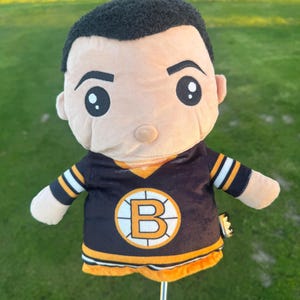 Pode incluir: Uma capa de pelúcia para a cabeça do taco de golfe em forma de personagem de desenho animado com cabelo preto e rosto bronzeado. O personagem usa uma camisa preta e dourada com a letra "B" na frente, representando o Boston Bruins.