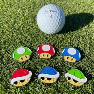 May include: A white Titleist golf ball with the number 4 rests on green grass, alongside six colourful mushroom and turtle shell golf ball markers. The markers are red, green, and blue with yellow and white accents.