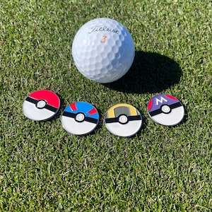 May include: A white Titleist golf ball with the number 3 and four colorful golf ball markers. The markers are designed with red, blue, yellow, and purple accents, resembling popular cartoon characters. The items are on a green grass surface.