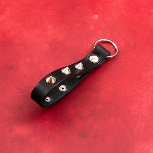 Peut inclure: Un porte-clés en cuir noir avec des clous pyramidaux argentés et un anneau argenté. Le porte-clés est sur un fond rouge.