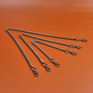 Puede incluir: Cadenas de metal plateado con cierres de mosquetón de diferentes longitudes, dispuestas en diagonal sobre un fondo naranja. Estas cadenas son probablemente para un bolso de mano u otro accesorio.