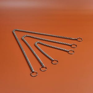 May include: Five silver metal chains with circular rings at the ends are arranged on an orange background. The chains vary in length, with the longest at the top and the shortest at the bottom. The chains are made of small, interlocking links.