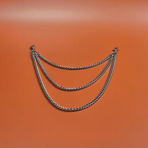 Pu&ograve; includere: Accessorio a catena argentato a tre livelli con chiusure a moschettone. Le catene sono distanziate uniformemente e pendono in una curva delicata su uno sfondo arancione. Le catene sono realizzate con piccole maglie.