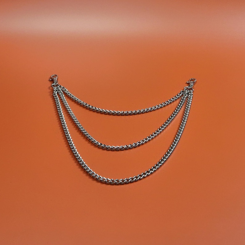 Pu&ograve; includere: Accessorio a catena argentato a tre livelli con chiusure a moschettone. Le catene sono distanziate uniformemente e pendono in una curva delicata su uno sfondo arancione. Le catene sono realizzate con piccole maglie.