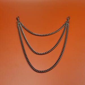 May include: A silver-coloured, three-tiered chain accessory with lobster claw clasps. The chains are connected at the top and hang down in a curved, symmetrical design. The background is a solid orange colour.