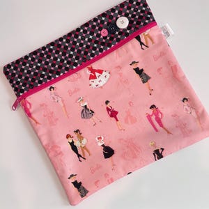 Puede incluir: Bolsa con cremallera de tela rosa con una parte superior de lunares negros y rosas. La bolsa est&aacute; decorada con ilustraciones de mujeres con vestidos y la palabra "Barbie". La bolsa se cierra con tres botones.