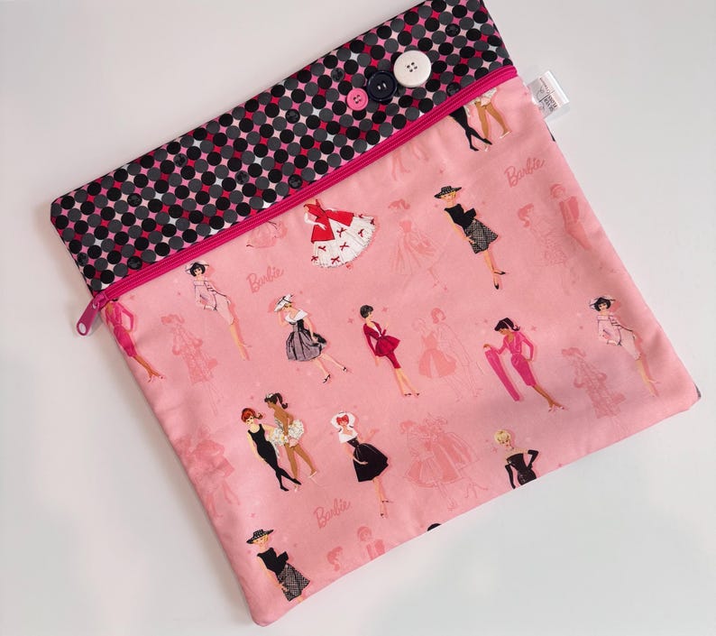 Puede incluir: Bolsa con cremallera de tela rosa con una parte superior de lunares negros y rosas. La bolsa est&aacute; decorada con ilustraciones de mujeres con vestidos y la palabra "Barbie". La bolsa se cierra con tres botones.