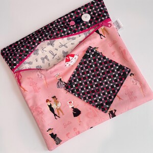 Puede incluir: Una bolsa de tela rosa y un posavasos cuadrado a juego con un estampado de mu&ntilde;eca Barbie. La bolsa tiene un estampado de lunares negros y grises en la solapa superior y un cierre de cremallera. El posavasos tiene el mismo estampado de lunares. Se ven botones y una cremallera.