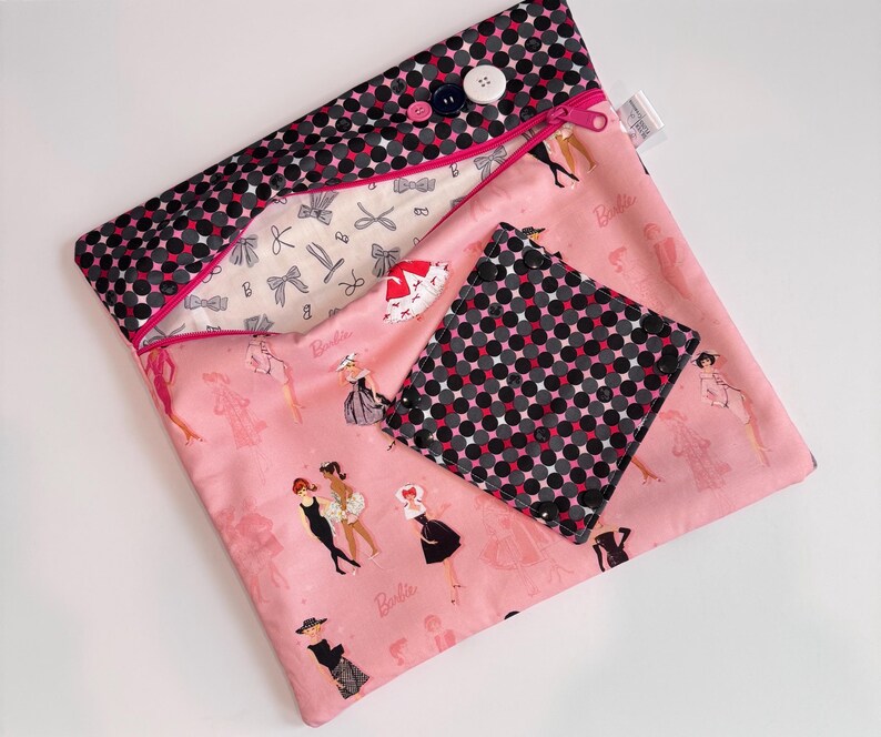 Puede incluir: Una bolsa de tela rosa y un posavasos cuadrado a juego con un estampado de mu&ntilde;eca Barbie. La bolsa tiene un estampado de lunares negros y grises en la solapa superior y un cierre de cremallera. El posavasos tiene el mismo estampado de lunares. Se ven botones y una cremallera.