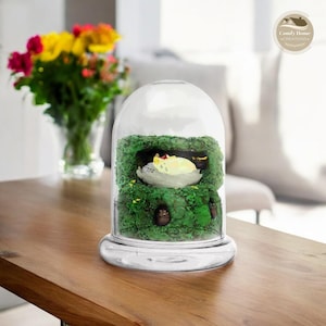 May include: A glass cloche with a clear base and dome, showcasing a miniature scene. Inside, a green mossy landscape houses a sleeping, light-yellow creature. The scene is set on a wooden table, with a vase of flowers in the background.