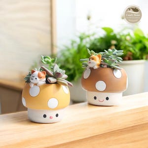 以下が含まれることがあります： かわいい動物の顔が付いたセラミック製のきのこ型のプランター2つ。左側のプランターは、白い水玉模様の黄色いきのこの帽子をかぶっており、寝ている猫の置物があります。右側のプランターは、白い水玉模様の茶色のきのこの帽子をかぶっており、小さな犬の置物があります。どちらのプランターにも、緑色の多肉植物が植えられています。