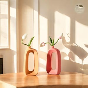 May include: Two ceramic vases with a unique, modern design. The vase on the left is orange and the vase on the right is pink. Both vases have a large, oval opening in the center. The vases are filled with artificial flowers.