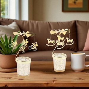 May include: Two decorative candle holders with gold-colored carousel-themed toppers. One features figures riding bicycles, the other has rocking horse figures. The candle holders have a textured, speckled glass design and sit on a wooden table.