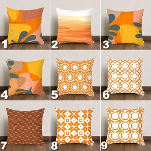 May include: Nine decorative throw pillows with various designs. Pillows feature abstract art, geometric patterns, and landscape scenes in shades of orange, yellow, and white. The pillows are square and rest on a wooden floor.