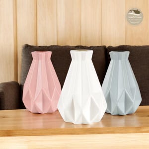 Può includere: Tre vasi geometrici in ceramica rosa, bianco e grigio. I vasi hanno un design a carta piegata e sono disposti su un tavolo di legno.