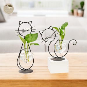 Op de afbeelding: Twee zwarte metalen plantenhouders in de vorm van een kat met glazen hartvormige vazen. Elke vaas bevat een kleine groene plant.