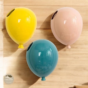 Könnte beinhalten: Drei Keramik-Ballon-Wandskulpturen in Gelb, Rosa und Türkis. Die Ballons sind glänzend und haben oben ein Loch.