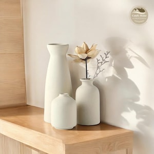 以下が含まれることがあります： 異なるサイズと形の白いセラミックの花瓶が3つあり、中央の花瓶にはドライフラワーの飾り付けがされています。花瓶は表面がテクスチャ加工されており、木製の棚に並べてあります。