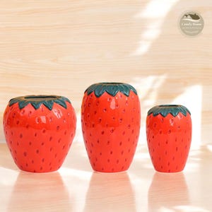 May include: Three red ceramic strawberry-shaped vases with green leaves. The vases are different sizes and are arranged in a row on a light wood surface.