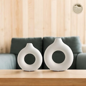 Può includere: Due vasi in ceramica bianca con un'apertura a forma di anello. I vasi sono su un tavolo di legno.