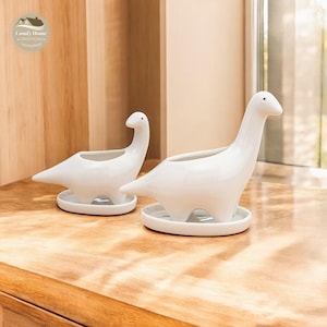 May include: Two white ceramic dinosaur planters. Each planter has a long neck and a small, round base. The planters are on a wooden surface. The planters are designed to hold small plants or succulents.