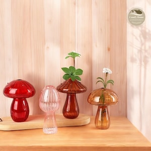 Puede incluir: Cuatro jarrones de vidrio con forma de hongo en rojo, rosa, marrón y ámbar. Los jarrones están dispuestos sobre una superficie de madera. Los jarrones están decorados con una textura acanalada. Los jarrones son perfectos para exhibir flores u otras plantas pequeñas.