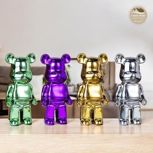 May include: Four glossy bear figurines in green, purple, gold, and silver. Each bear has a rounded head with small ears, a rectangular body, and short legs. The figurines are displayed on a wooden surface.
