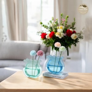 Peut inclure: Deux vases en plastique transparent en forme de bouton de fleur, avec une teinte bleu clair et vert clair. Chaque vase contient une seule tige avec deux petites fleurs en pompon de couleur pastel. Les vases sont posés sur un livre intitulé "Made & Crafted".