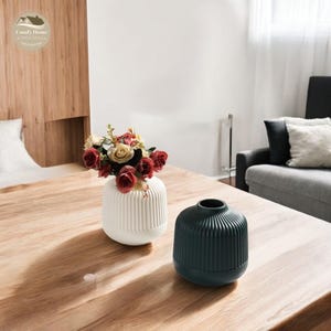 Könnte beinhalten: Zwei Keramikvasen, eine weiße und eine dunkelgrüne, mit einem gerippten Design. Die weiße Vase ist mit einem Strauß roter und gelber Rosen gefüllt. Die Vasen stehen auf einem Holztisch.