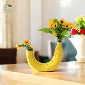 May include: A yellow ceramic banana-shaped vase with a black outline. The vase is filled with a bouquet of yellow sunflowers.