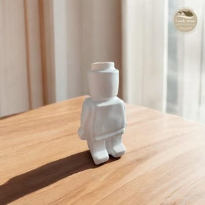 May include: White ceramic figurine shaped like a person, resembling a Lego figure, with a hollow top for holding flowers or other small items.