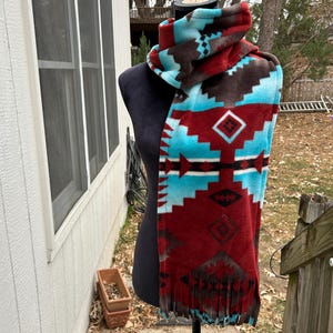 May include: A fleece scarf with a Southwestern pattern in shades of red, turquoise, and brown. The scarf has a fringed edge and is displayed on a black mannequin. The design features geometric shapes and patterns.