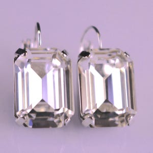 May include: A pair of clear, rectangular crystal earrings with silver-colored hooks. The crystals have a faceted design, reflecting light. The earrings are set against a soft purple background, showcasing their sparkle.