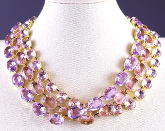 May include: A three-strand necklace with oval gemstones in shades of purple and pink. The gemstones are set in gold-tone metal. The necklace is displayed on a white bust.